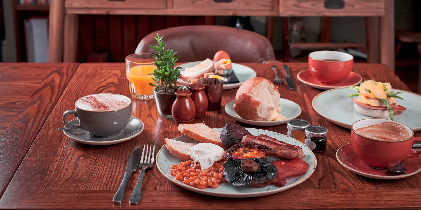 A wooden table with various breakfast related dishes on it. There is also tea, orange juice, jam and cutlery on the table