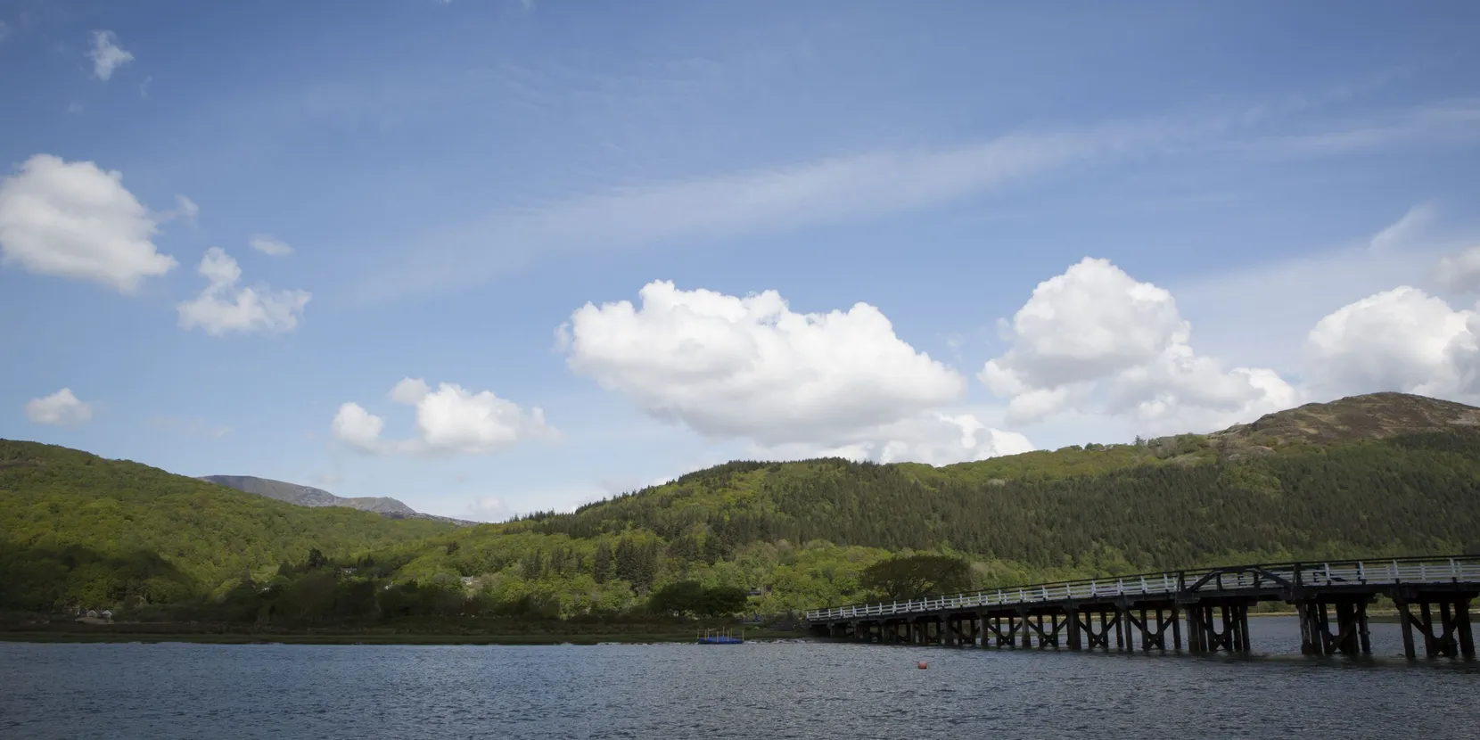 A landscape shot of mountains and water in front of it. There is also a bridge on the water.