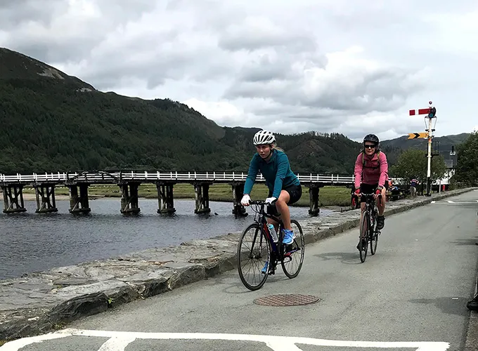 Two people cycling. Next to them is a river, bridge and hills in the background