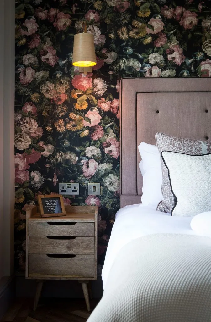A room with a part of the bed showing. There is also a small chest of drawers next to it and a light above them both.