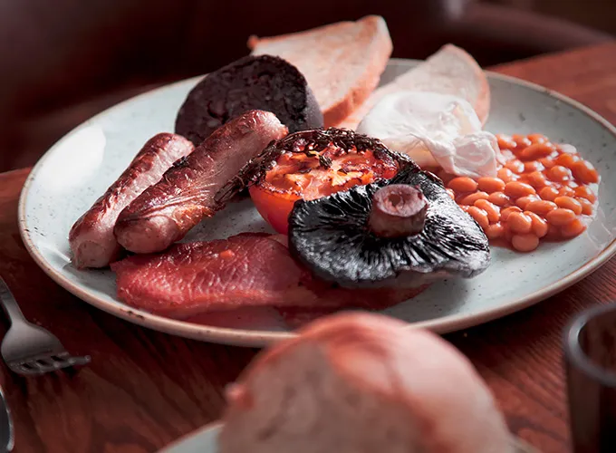 English Breakfast on a white plate. There is beans, sausage, bacon, tomato, toast and other items associated with an English breakfast on the plate