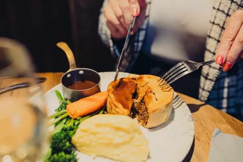 Someone cutting into a pie. Next to the pie is mash potatoes, carrots, broccoli and gravy