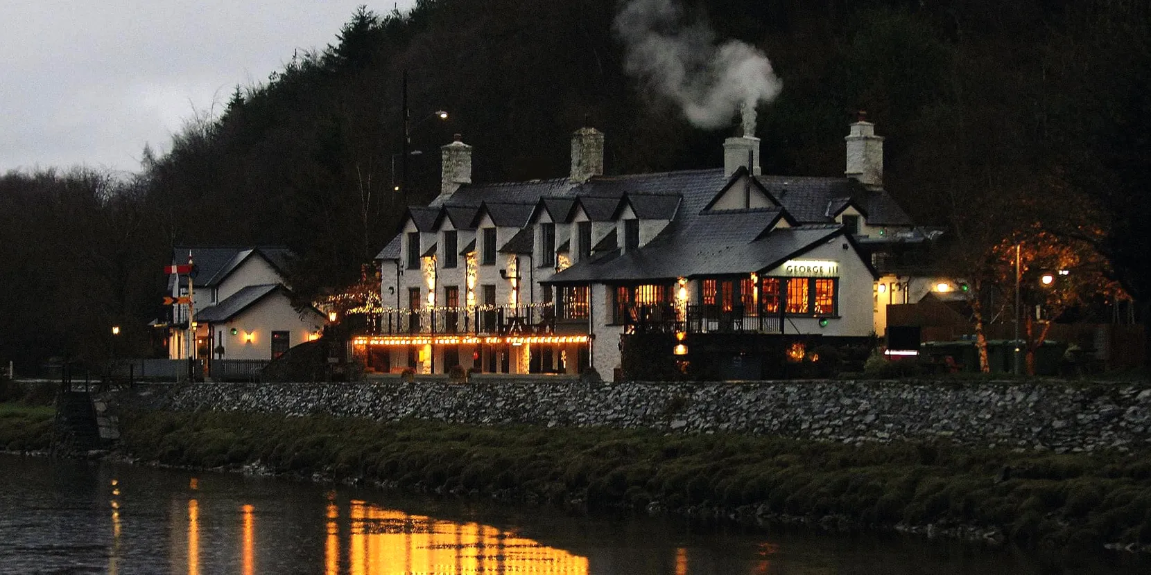 A landscape shot of the George pub. It is becoming night and there are various lights on. In front of the pub is the river.